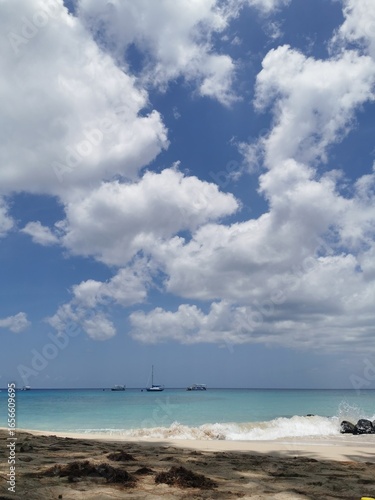 Dynamic tropical beach scene with waves breaking on the shore, dark sand, and several boats anchored in the turquoise ocean under a cloudy sky