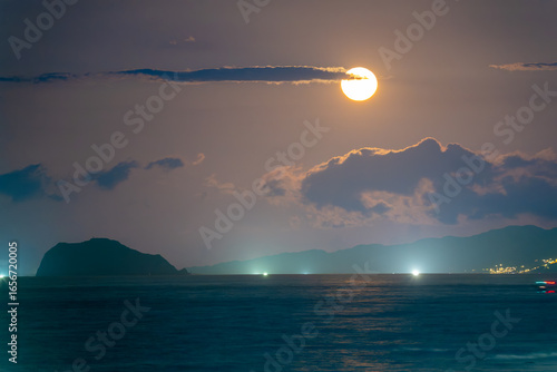 The full moon ascends from behind the clouds over the Shimen coast in New Taipei City, Taiwan, illuminating Xiao Jilong Yu (Little Keelung Island) and busy fishing boats at night.