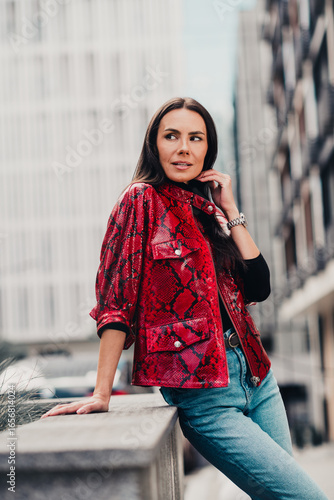 Glamorous brunette in red patterned jacket posing outdoors in urban city environment during a casual daylight stroll, vertical photo