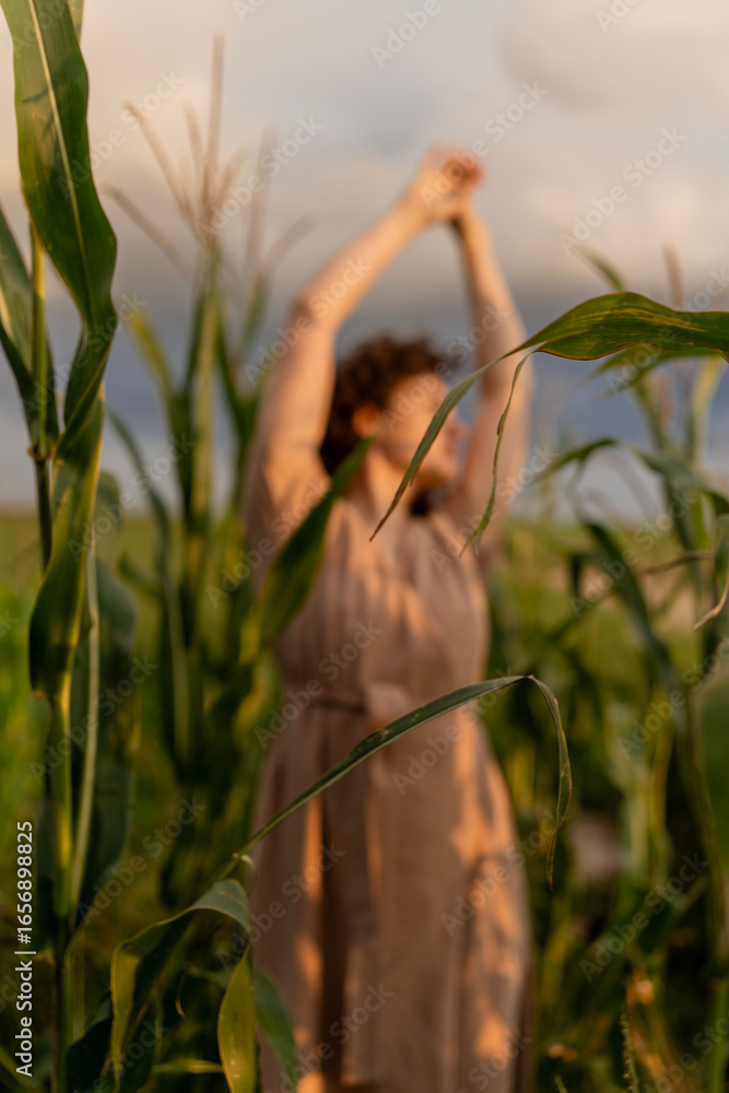 Obraz premium The concept of love for nature. Young woman with curly hair stretching in a cornfield during sunset, wearing a beige dress with a blurred background.