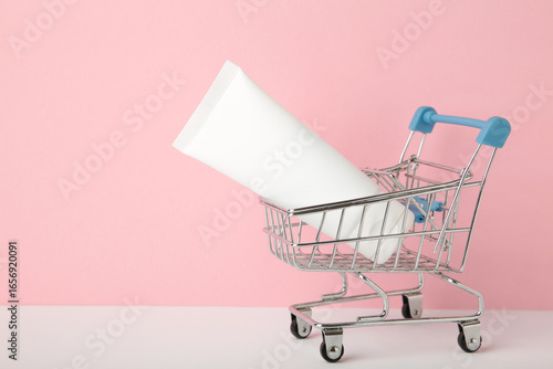 A mini shopping cart holds a white tube product on pink background. Perfect for concepts of retail and personal care products.