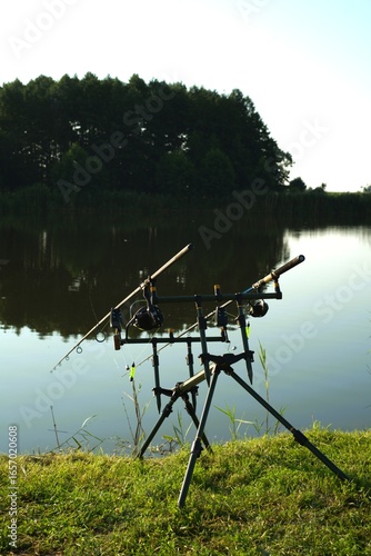 A fishing setup on a tripod stand placed by a peaceful lake. The image shows fishing rods with reels, prepared for fishing. The calm water and lush greenery create a relaxing outdoor scene.