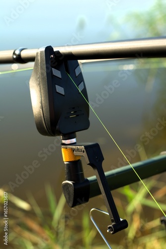 Close-up of a fishing rod with a line connected to an alarm system. The photo shows the reel, line, and electronic bite indicator in a peaceful outdoor fishing environment near a calm lake.
