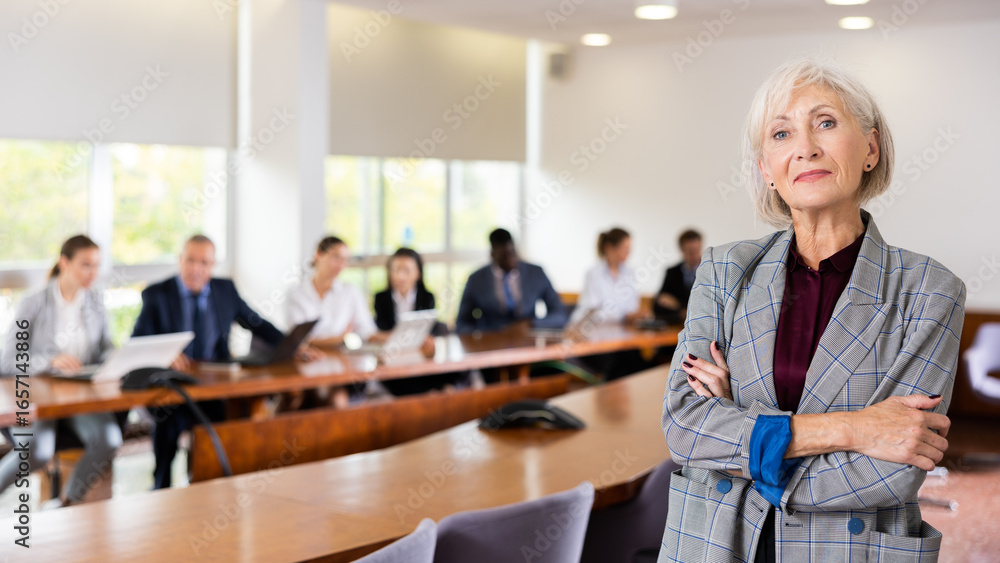 Obraz premium Smiling classy elegant senior mature white businesswoman on foreground posing at camera with arms crossed and office employees at back