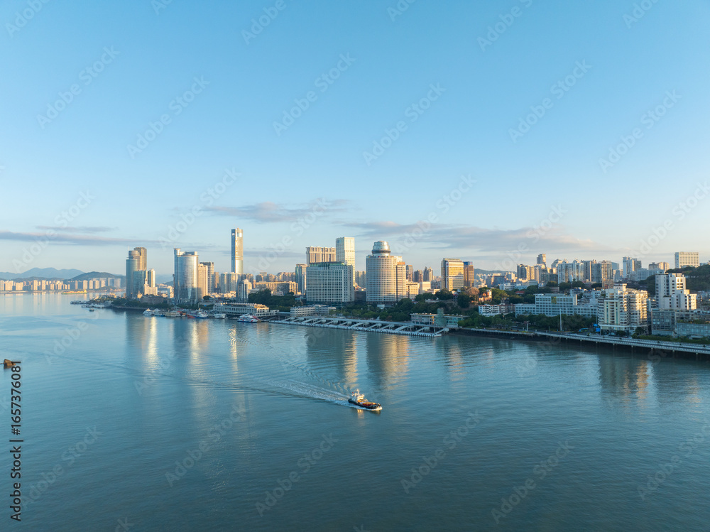 Naklejka premium Coastal Xiamen China cityscape reflects morning light on tranquil water as boats glide past modern waterfront buildings.