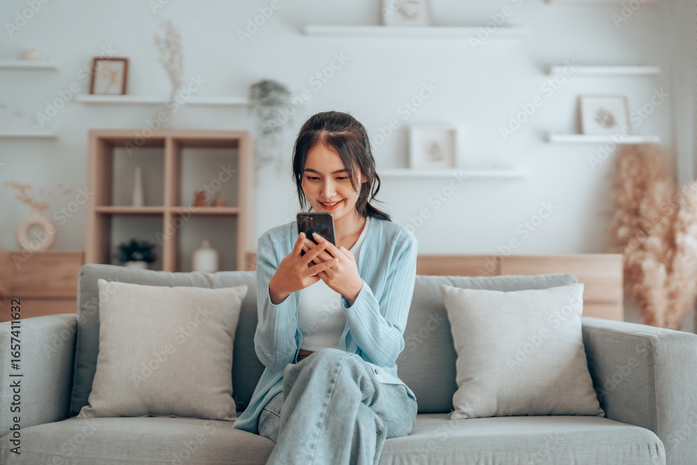 Smiling pretty young Asian woman sitting on cozy couch, using funny mobile apps in living room. Woman at Home, Doing Online Shopping, Messaging Friends, Posting on Social Media Smiling pretty young Asian woman sitting on cozy couch, using funny mobile apps in living room. Woman at Home, Doing Online Shopping, Messaging Friends, Posting on Social Media