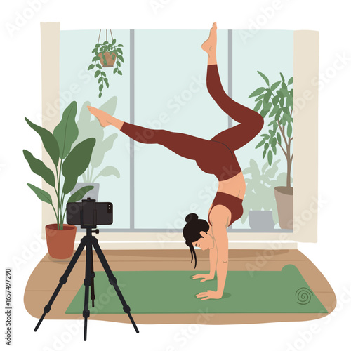 A woman is practicing yoga in a handstand position on a yoga mat in a room with large windows. She is dressed in a maroon sports bra and leggings