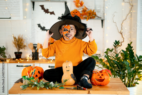 Halloween, a child boy in a festive costume and makeup is preparing for Halloween at home in the kitchen by coloring and carving pumpkins and scaring everyone