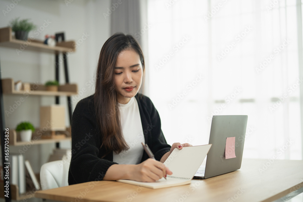 Young woman writing notes in a notebook while sitting at a desk with a laptop, minimalistic workspace, natural light, focus on productivity and organization Young woman writing notes in a notebook while sitting at a desk with a laptop, minimalistic workspace, natural light, focus on productivity and organization