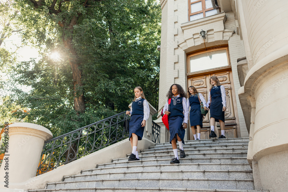 Fototapeta premium Group of schoolgirls in uniform walking down stone stairs with confidence. Concept of schoolwear fashion, education promotion, teen friendship and campus lifestyle.