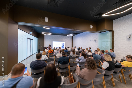People attending a professional presentation in a modern conference room with a speaker presenting on a screen.