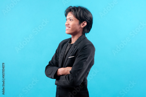 A young man in a black suit standing in side profile with folded arms and a cheerful smile, representing confidence, optimism, and modern professional style on blue background.