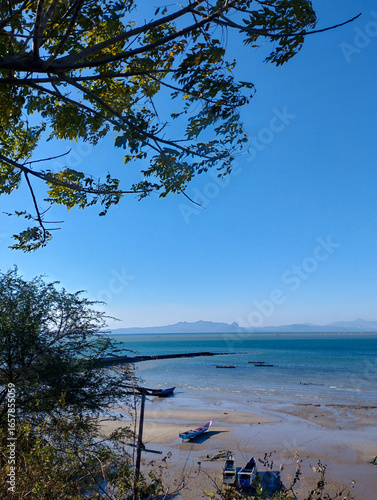 Scenic coastal view with small fishing boats on a sandy shore, calm turquoise sea, and distant mountains under a clear blue sky