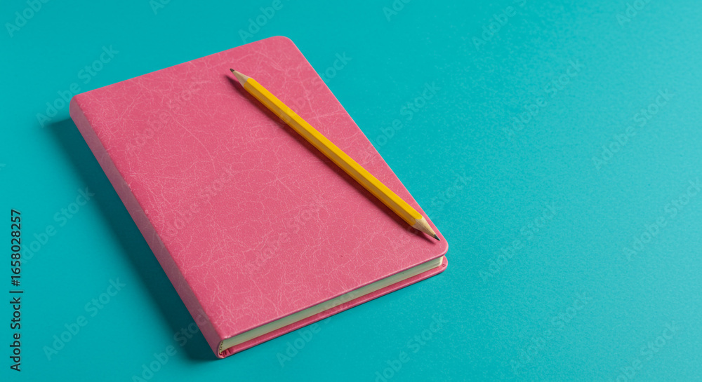 Fototapeta premium A pink notebook with a yellow pencil lying diagonally across it on a blue background flat lay