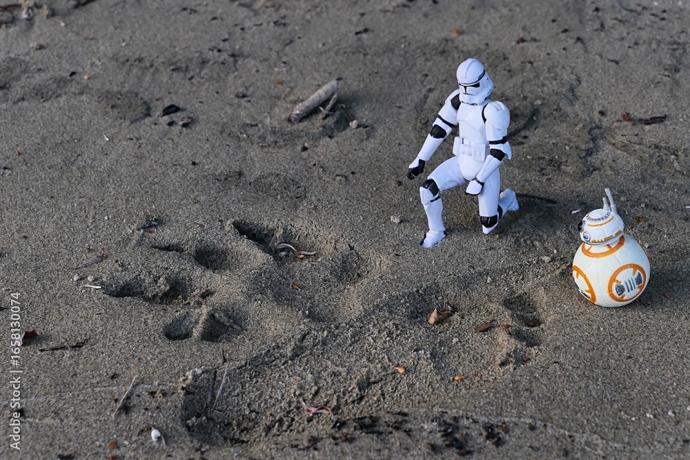 Fototapeta premium Figures of Star Wars clone trooper and ball shaped astromech droid BB8 examining tracks of a dog and little child in sand on shoreline of Danube river, southern Slovakia. 