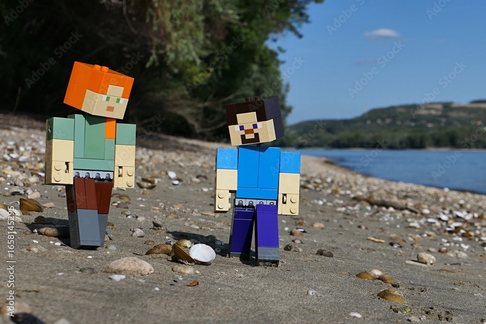 Obraz premium LEGO Minecraft figures of Steve and Alex, searching for clams and stones on sandy river Danube bank, borderline between Slovakia and Hungary. Forested opposite bank and blue skies visible.