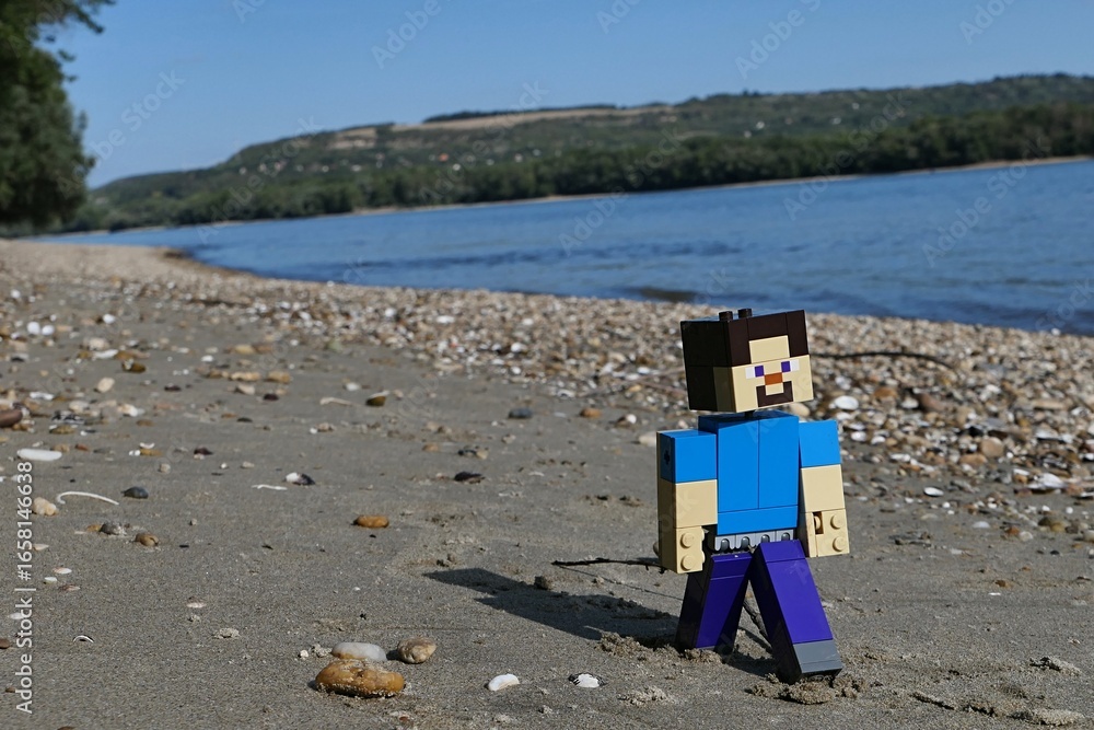 Obraz premium LEGO Minecraft action figure of smiling Steve walking on sandy bank of Danube river on borderline between Slovakia and Hungary. Forested opposite bank visible, blue skies, summer afternoon sunshine.