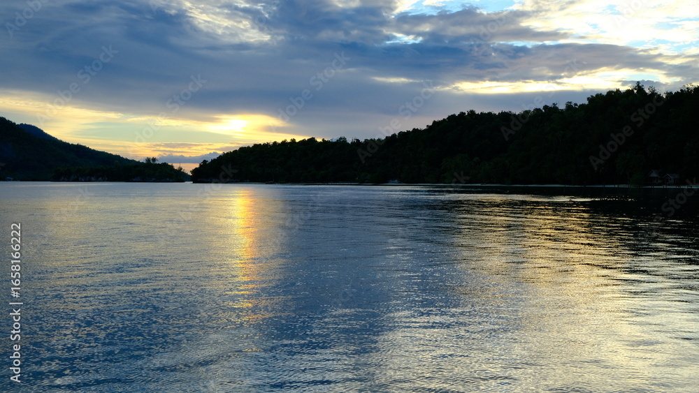 Fototapeta premium Colorful sunset over tropical islands with orange and purple sky reflecting on the calm sea in Raja Ampat, Indonesia symbol of travel and relaxation