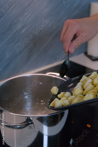 A person uses a spoon to slide raw potato gnocchi from a baking sheet into a pot of boiling water on a stove.