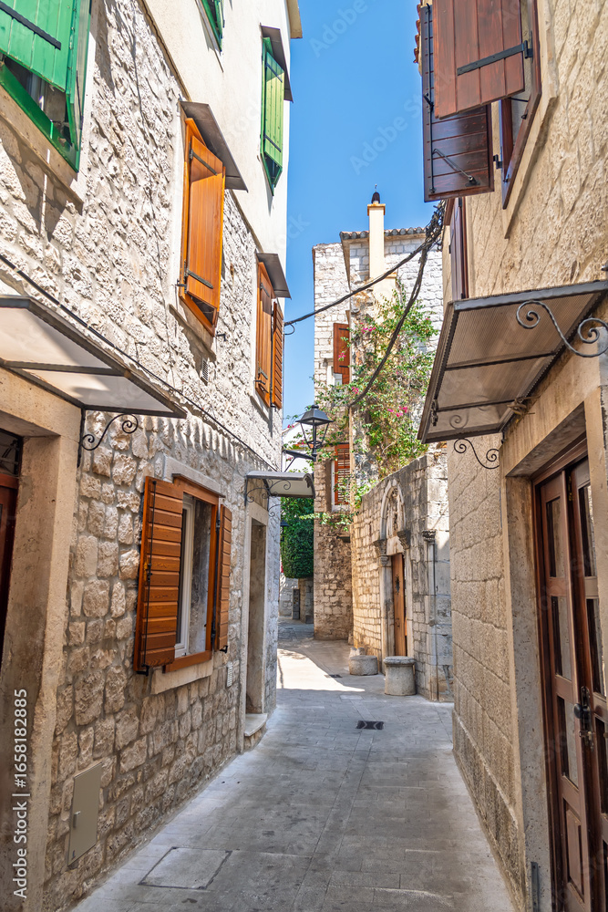 Fototapeta premium A charming narrow alley in a historic Mediterranean old town, with traditional stone houses and colorful wooden shutters on a sunny day with shadows and light playing on the ancient stone facades