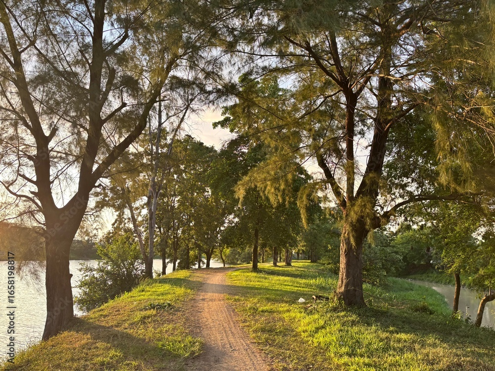 Naklejka premium Scenic dirt path beside a tranquil lake with lush trees, silhouetted against a golden sunset sky, with a distant power plant or factory on the horizon