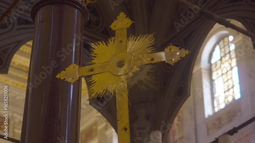 Golden processional cross with radiant sunburst design mounted on ornate column, displayed inside historic basilica with arched windows and decorative ceiling in background