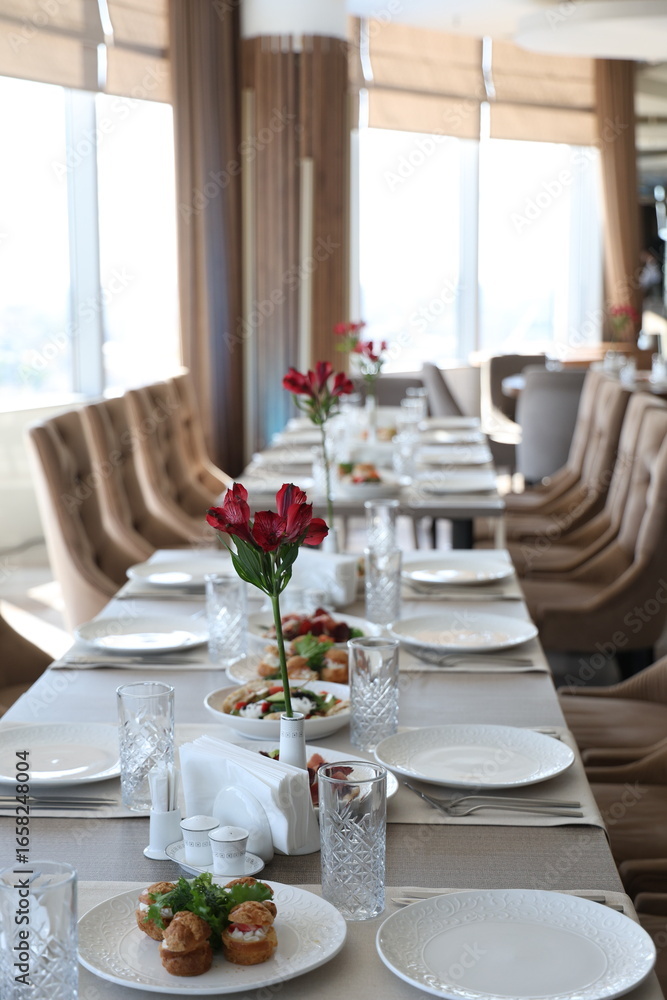 Fototapeta premium Vertical photo of formal dining table with appetizers and floral decor in sunlit room