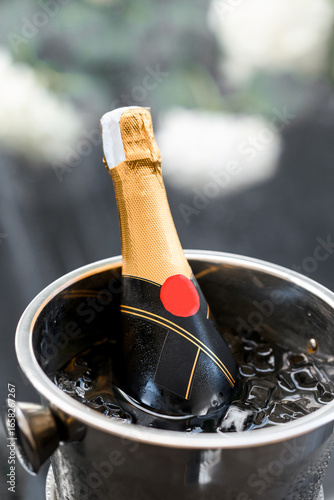 A bottle of sparkling wine with gold foil and black label resting in a metal ice bucket