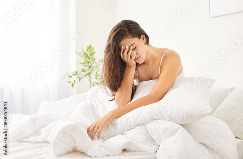 Foto Upset young Caucasian woman sitting in bed with pillows and blanket, leaning her head on her hand after bad night sleep