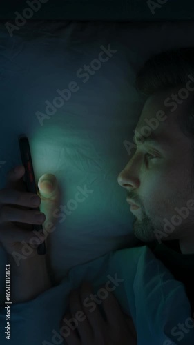 A tired man in bed scrolls endlessly on his mobile phone, struggling with insomnia, anxiety and overthinking as he watches social media late into the night. Smartphone dependence. vertical video