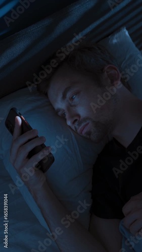Tired young man lies in bed at night, staring at his smartphone screen, doom scrolling through social media, unable to sleep struggling with sleep disorder or distraction by overthinking. 