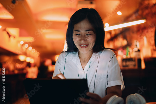 Schilderij op canvas Asian woman draws on digital tablet in ambient cafe light, illustrating modern creative process, mobile artistry and seamless tech integration into freelance workflow