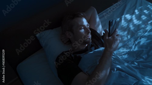 An attractive young man uses his phone late at night in bed, scrolling through his social media feed. Space for text