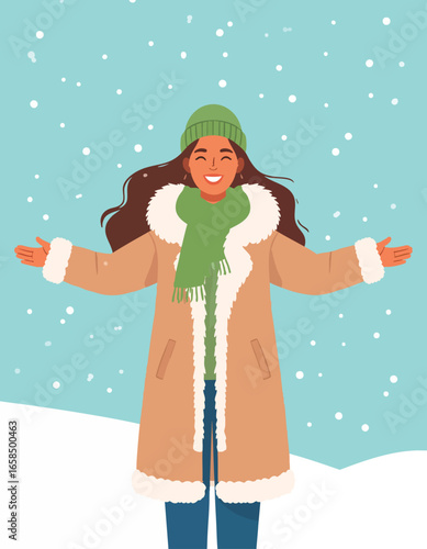 A smiling woman in winter clothing stands with open arms, enjoying falling snow.