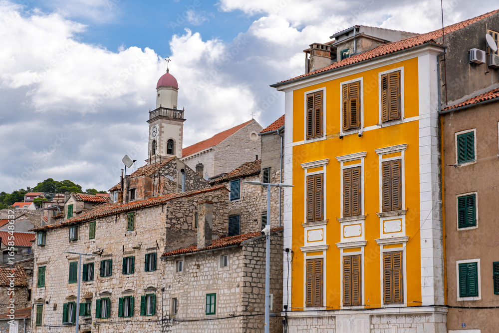 Fototapeta premium Cityscape of Sibenik, Croatia, featuring traditional terracotta roofs, wooden shutters, and a bell tower. Facade of a vibrant yellow building and the Church of St. Francis in the historic center