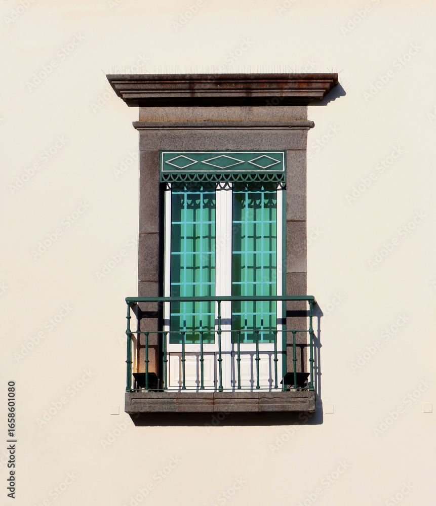 Obraz premium Classic window with green shutters and small balcony on a beige wall. Mediterranean architectural detail with iron railing and stone frame.