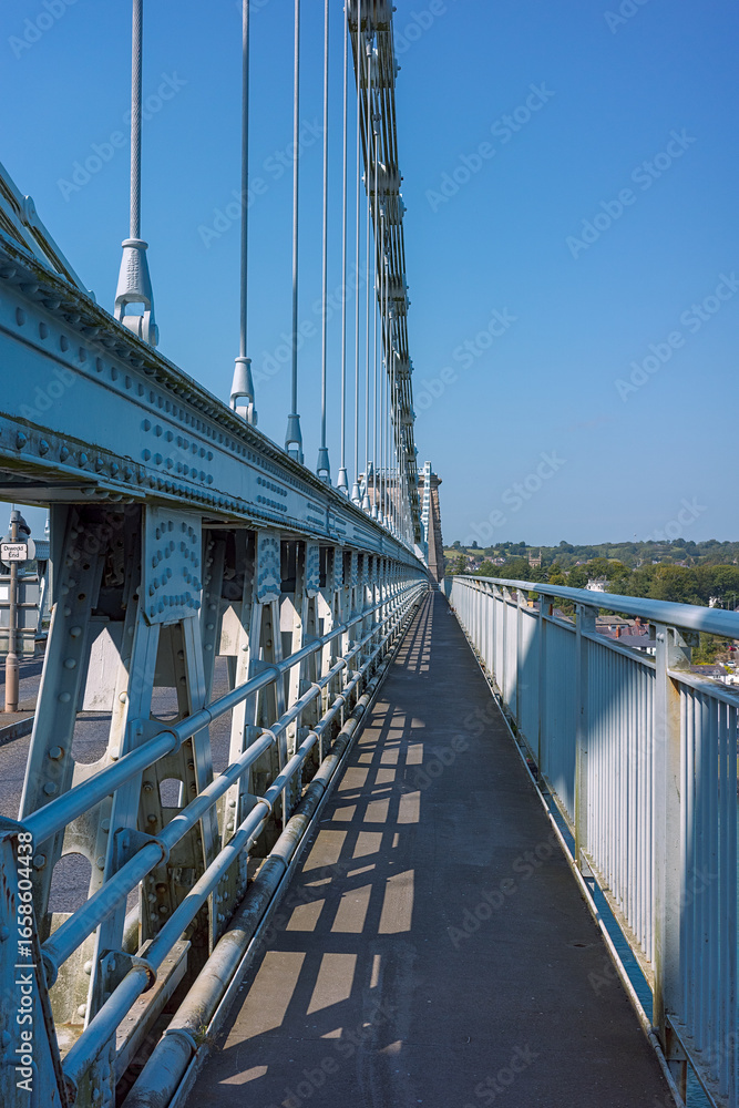 Naklejka premium Menai Bridge Anglesey North Wales. Elevated footpath. Perspective of a historic suspension bridge under a clear blue sky.