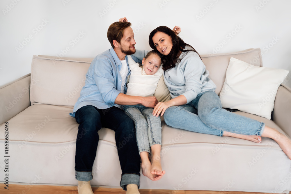Family of three sits close together on beige couch, smiling toward camera. Hidden tablet emphasizes quiet presence of tech in serene domestic life and modern digital comfort zones. Family of three sits close together on beige couch, smiling toward camera. Hidden tablet emphasizes quiet presence of tech in serene domestic life and modern digital comfort zones.