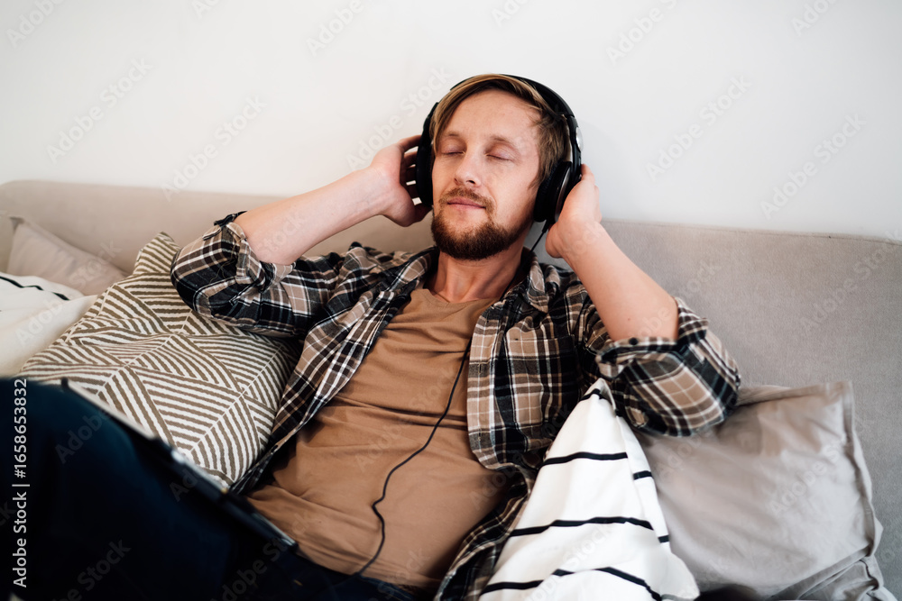 Obraz premium Man enjoying immersive digital moment, eyes closed with headphones on, resting on bed with tablet by side, symbolizing audio therapy, creative inspiration, and tech balance.