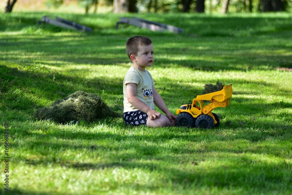 Fototapeta premium Little boy sitting on green lawn with yellow toy truck filled with grass. Summer outdoor play, childhood imagination and leisure in countryside nature, joyful and carefree moment.
