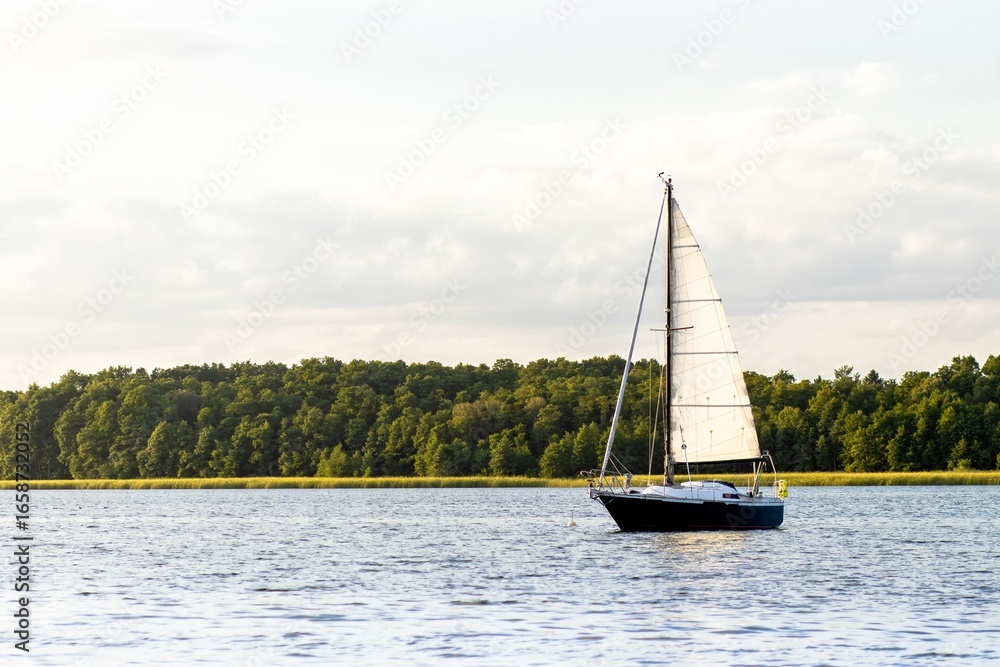 Obraz premium Sailboat gliding across calm waters, surrounded by lush green trees on the shoreline, under a bright sky with soft clouds, capturing the essence of peaceful sailing adventure