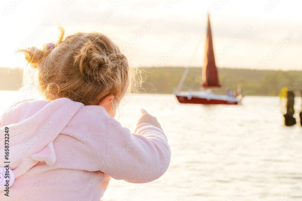 Fototapeta premium Young girl in pink hoodie points towards a sailboat on a shimmering lake during sunset, capturing a moment of wonder and exploration in a serene outdoor setting