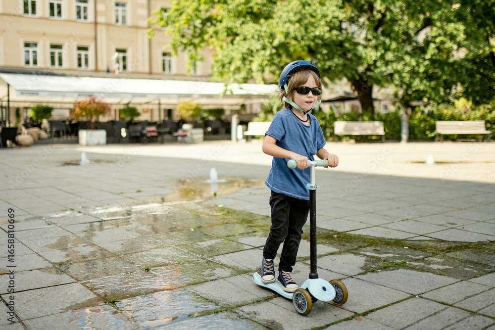 Fototapeta premium Young boy rides a scooter through a sunlit square in Vilnius, wearing a helmet and sunglasses, enjoying a warm day