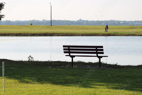 Peaceful Bench by the Lake With a Scenic View, Inviting Relaxation and Contemplation in the Afternoon Sun, Surrounded by Lush Greenery and Distant Walking Figures