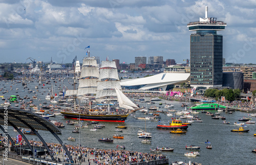 Canvas Print A vibrant view of Amsterdam Central Station with NS trains, trams, and a large crowd gathered along the IJ river during a maritime event featuring numerous boats and a tall sailing ship
