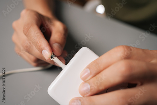 Fotografie Technician hands inspecting damaged charging cable, revealing connectivity risks from visible cable fraying and potential electrical safety concerns, sitting at desk in workshop