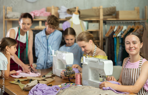 Bild auf Leinwand Engaged teenage schoolgirl enjoying group sewing class, stitching at machine in school tailoring studio while peers working with fabric and pattern under guidance of female instructor
