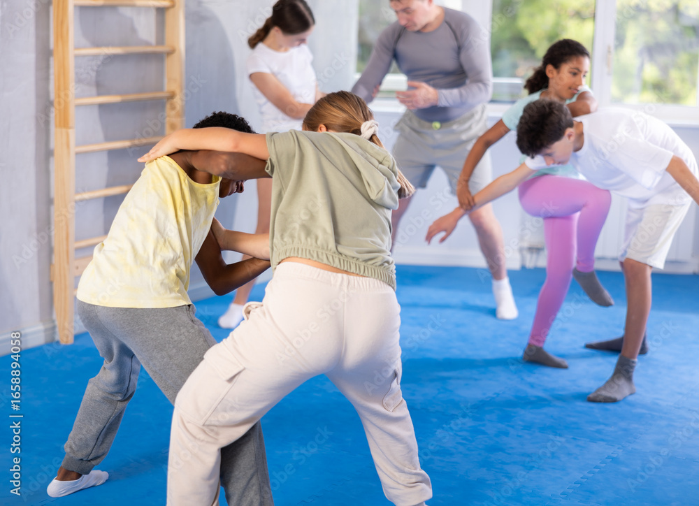 Fototapeta premium Girls and boys practicing in pair self-defence movements with male trainer supervision