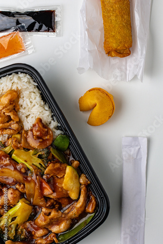 Flat lay of Asian takeout food featuring chicken stir fry with broccoli and peppers, white rice, fried egg roll, fortune cookie, and dipping sauces. Fast food concept on clean minimal background.