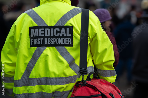 Close-up of a medical first responder wearing a bright fluorescent yellow coat with a grey reflective cross. The emergency EMT officer has a red first aid bag with a black strap over their shoulder. 